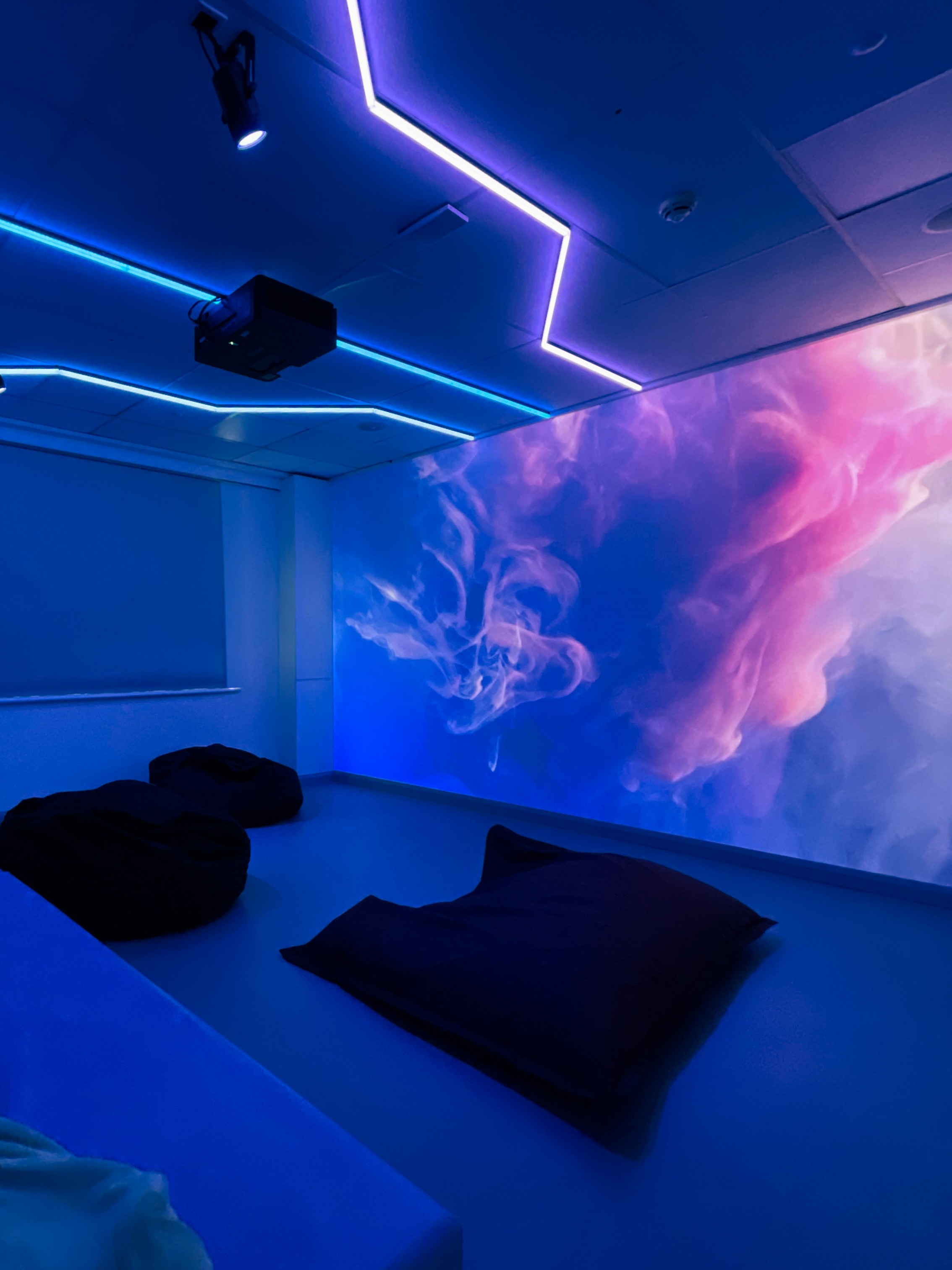 A room with ambient neon blue and purple lighting, featuring bean bags and a large wall display of swirling pink and blue clouds, creating a tranquil atmosphere.