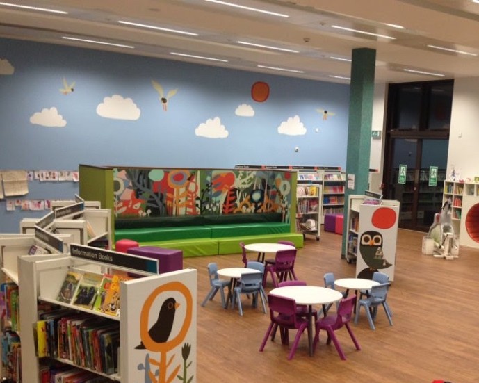 Children's library area with colorful wall art of clouds and birds. Bookshelves and small tables with chairs create a welcoming, playful atmosphere.