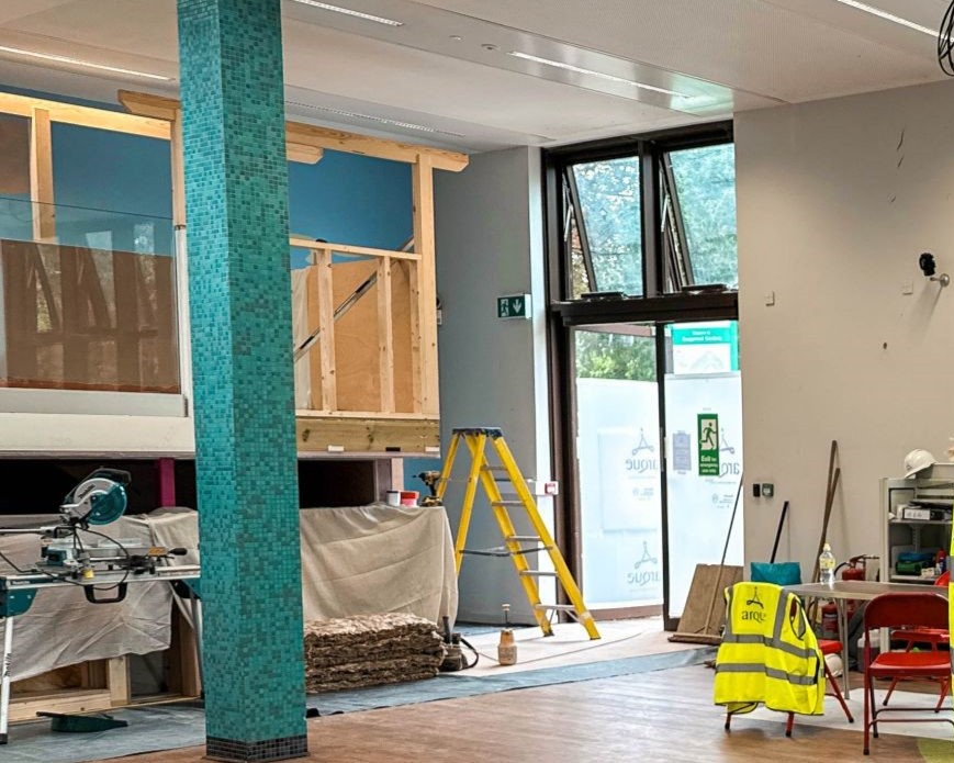Construction site with wooden structures, a yellow ladder, and tools scattered. Bright interior with large windows, a mosaic column, and a safety vest hung on a chair.