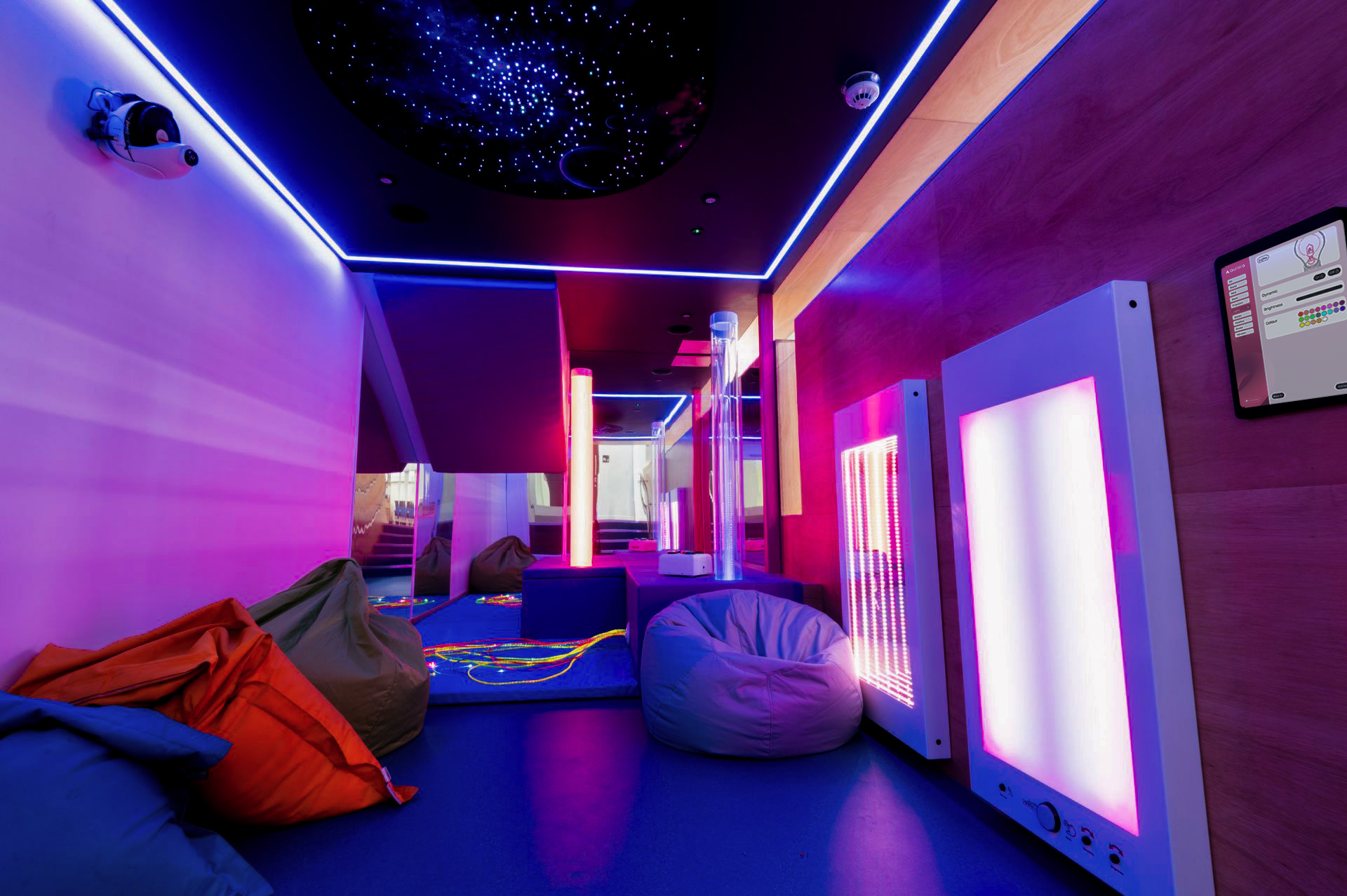 A futuristic neon-lit room dominated by vibrant purples, pinks, and blues. The walls and ceiling feature bright neon strip lighting, with a starfield effect on the ceiling. The space contains bean bag chairs in red and purple, illuminated panels with glowing edges, a tall central pole, and a blue floor. A monitor is visible on the right wall. The overall aesthetic is modern and gaming-focused with a cyberpunk vibe.