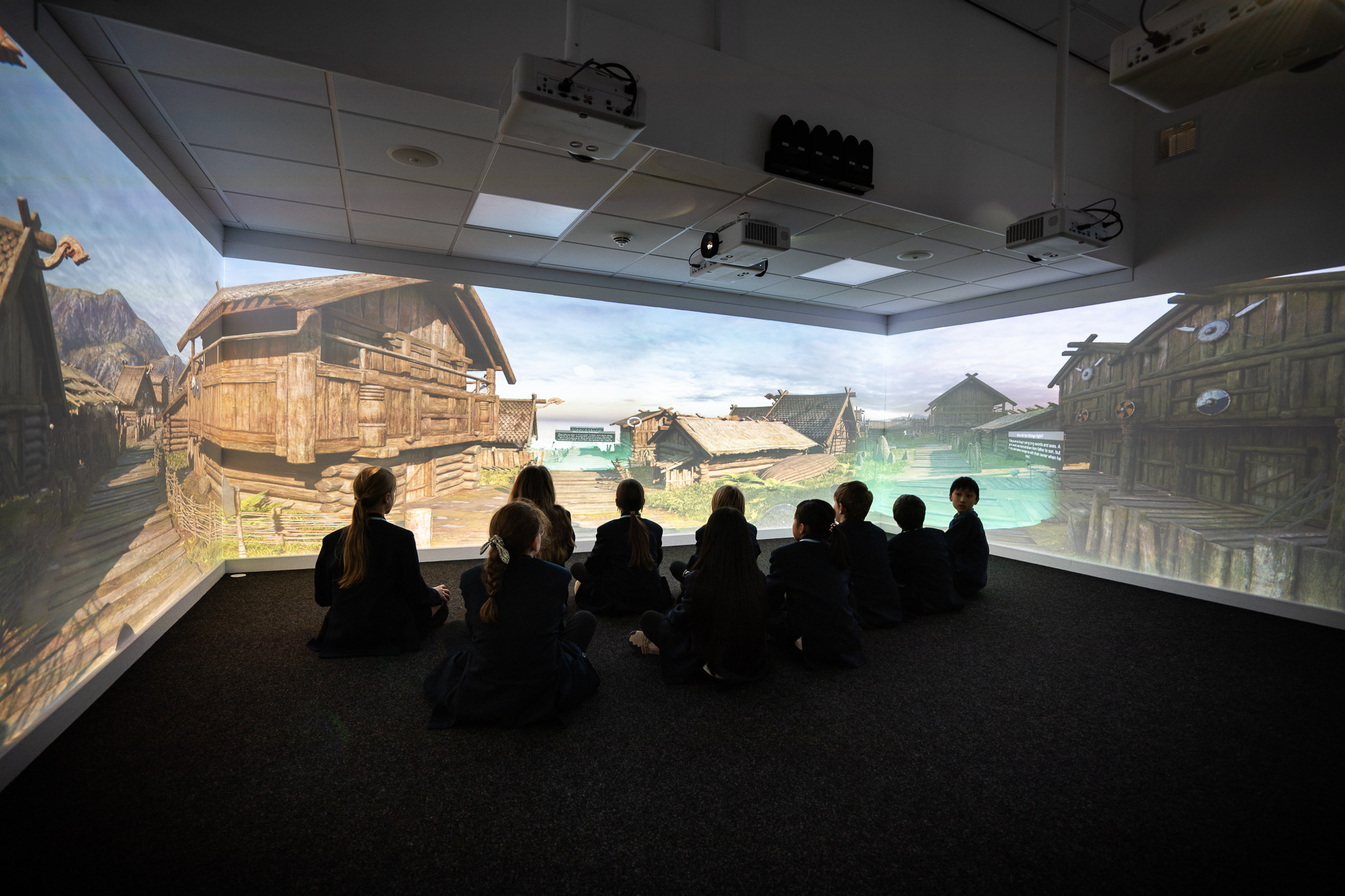 Children sit in a dark room facing a wrap-around screen displaying a virtual medieval village scene, creating an immersive learning environment.