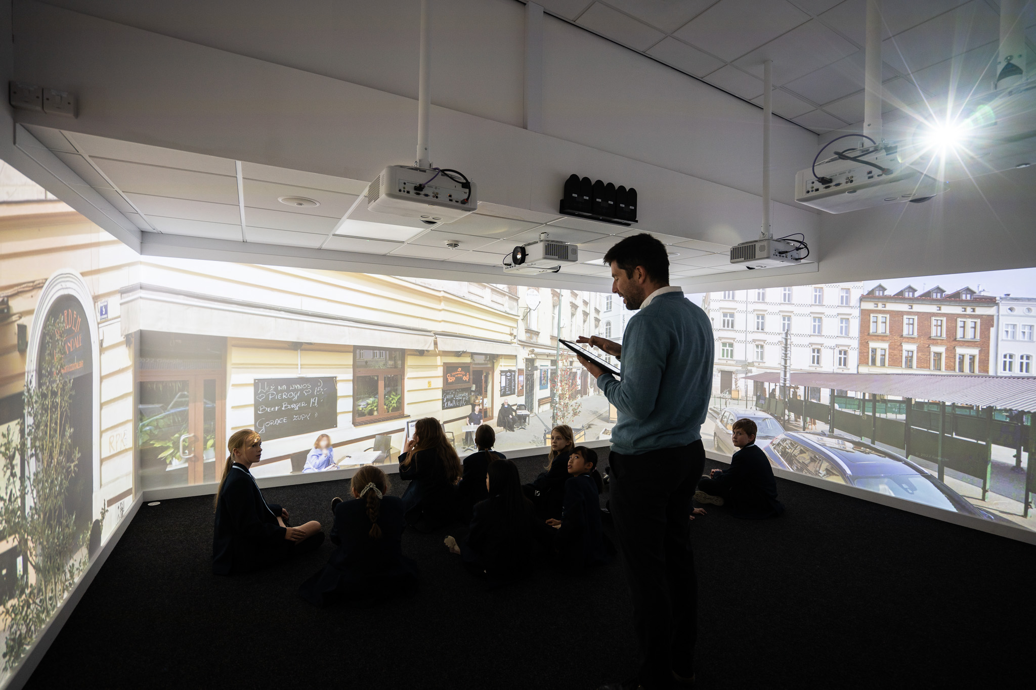 A group of students sits on the floor in a dark room with large projected images of a street scene. A teacher stands nearby, holding a tablet.