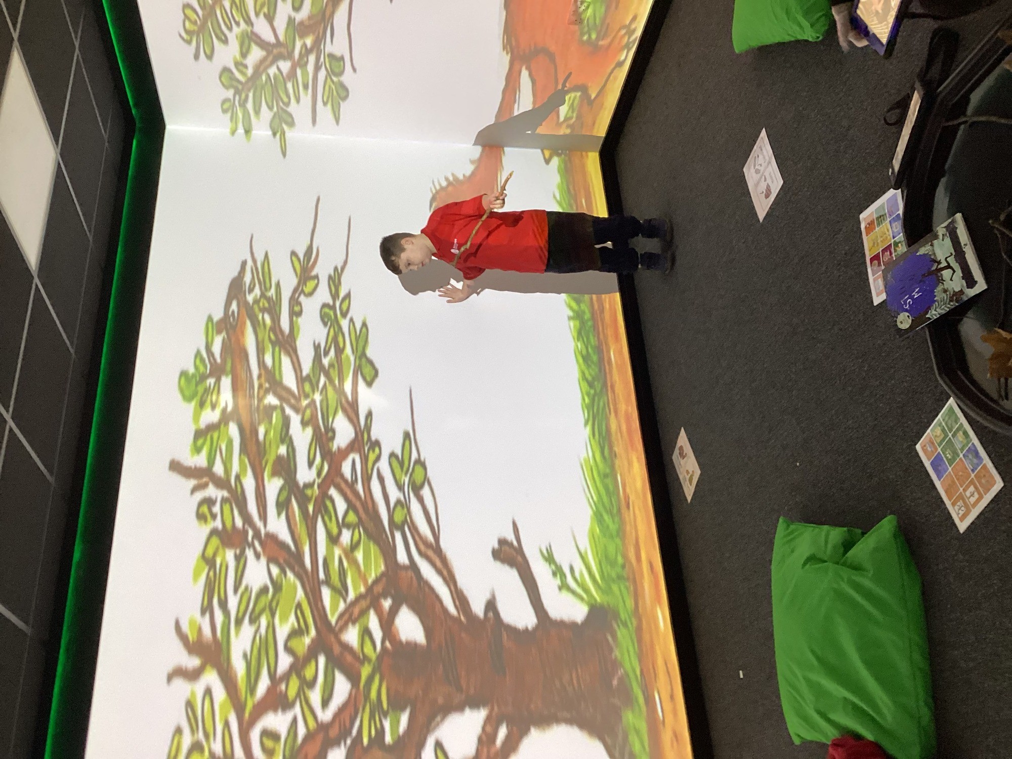 A person in a red shirt interacts with a large digital tree projected on a wall. Green beanbags and paper sheets are on the floor, creating an immersive environment.