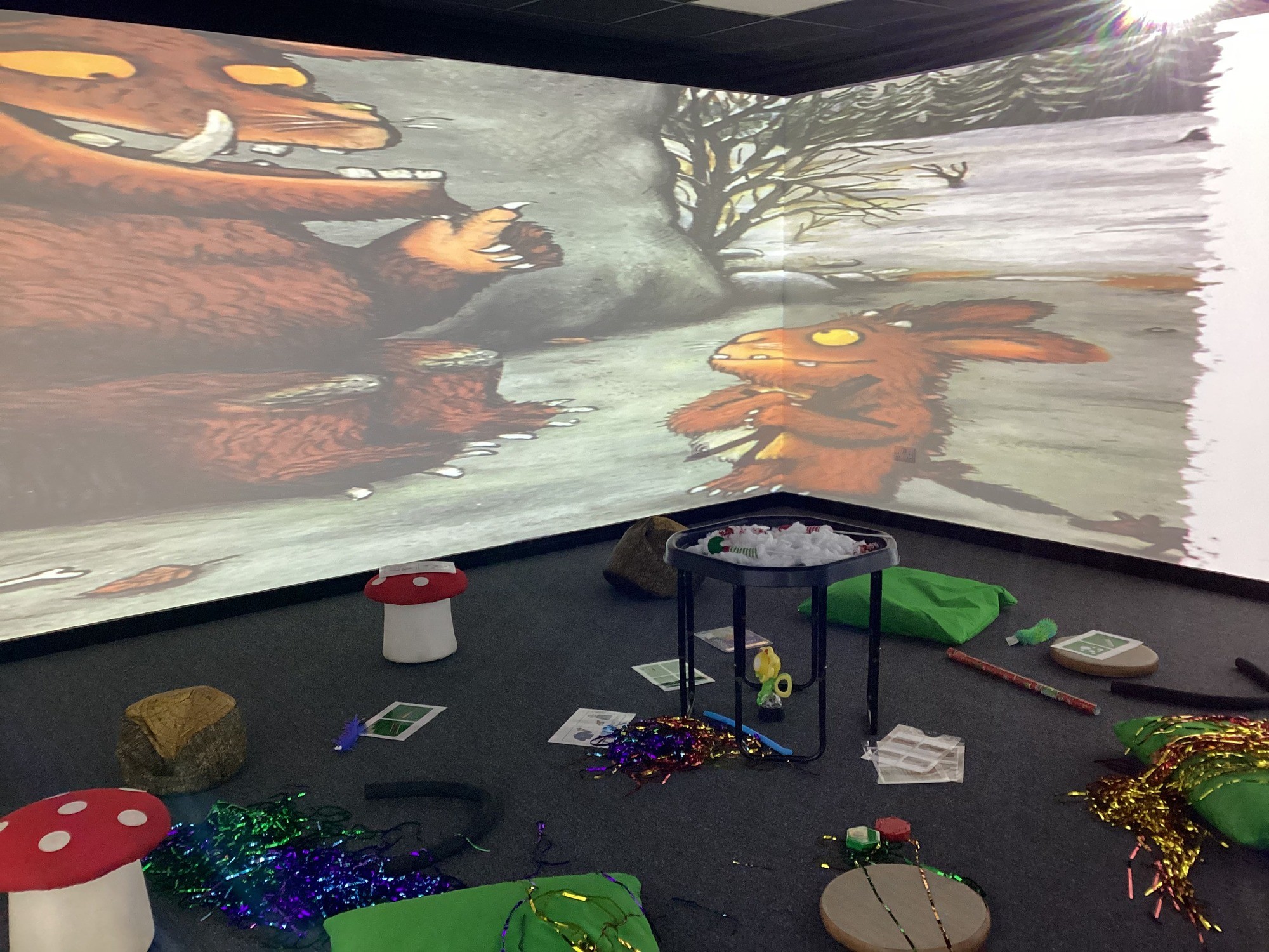 Room with Gruffalo-themed wall projection showing large creature and smaller creature in a snowy forest. Floor has toy mushrooms, tinsel, and activity papers, creating a playful, whimsical atmosphere.