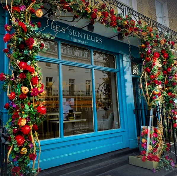 Storefront of a perfumery with blue walls adorned with vibrant red and pink flower garlands. Large windows reflect nearby buildings, creating an inviting, elegant atmosphere.