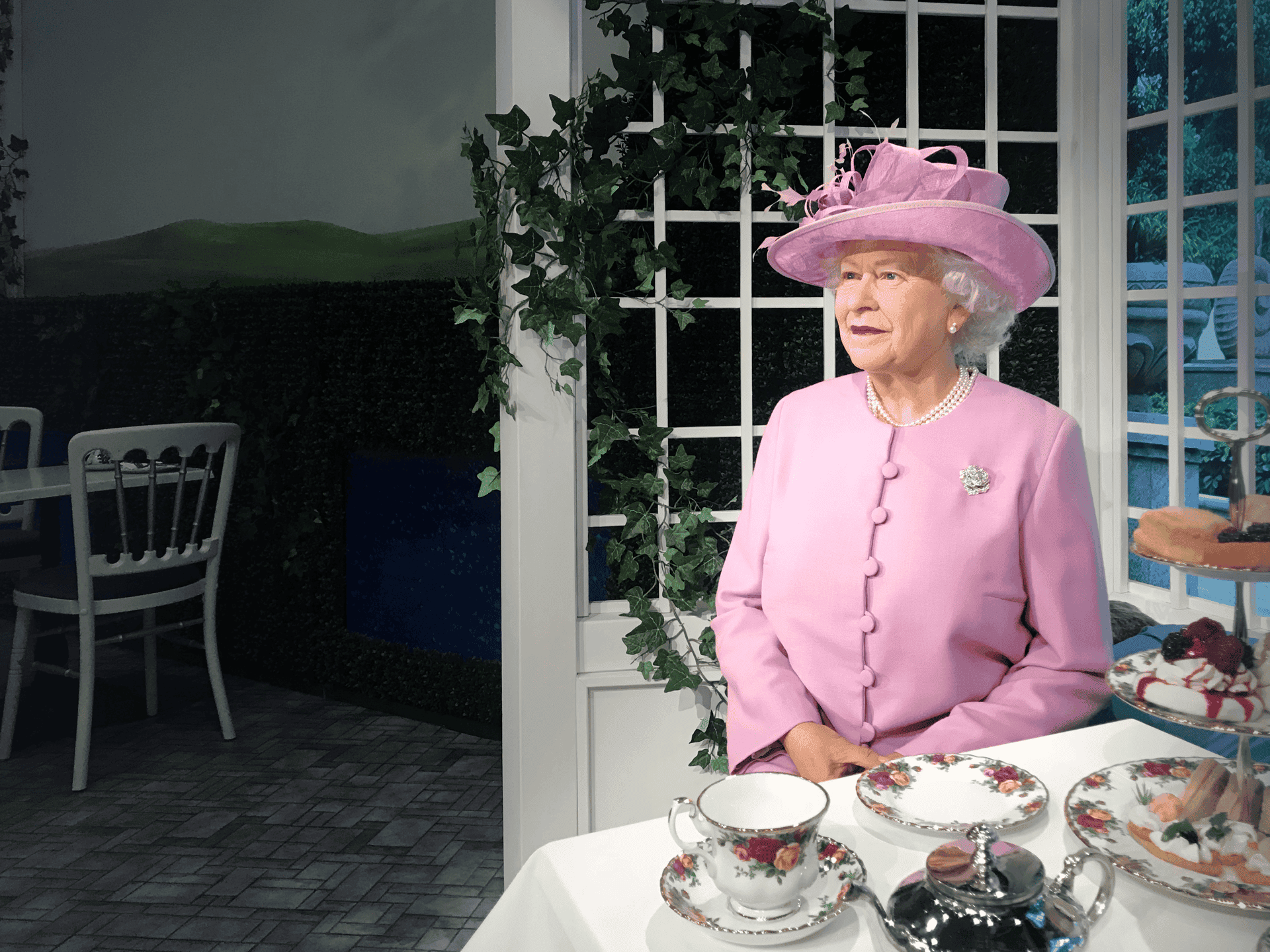 Wax figure of Queen Elizabeth II in a pink outfit and hat, sitting at a table with floral china, pastries, and tea. The setting is elegant, with ivy and a garden view.