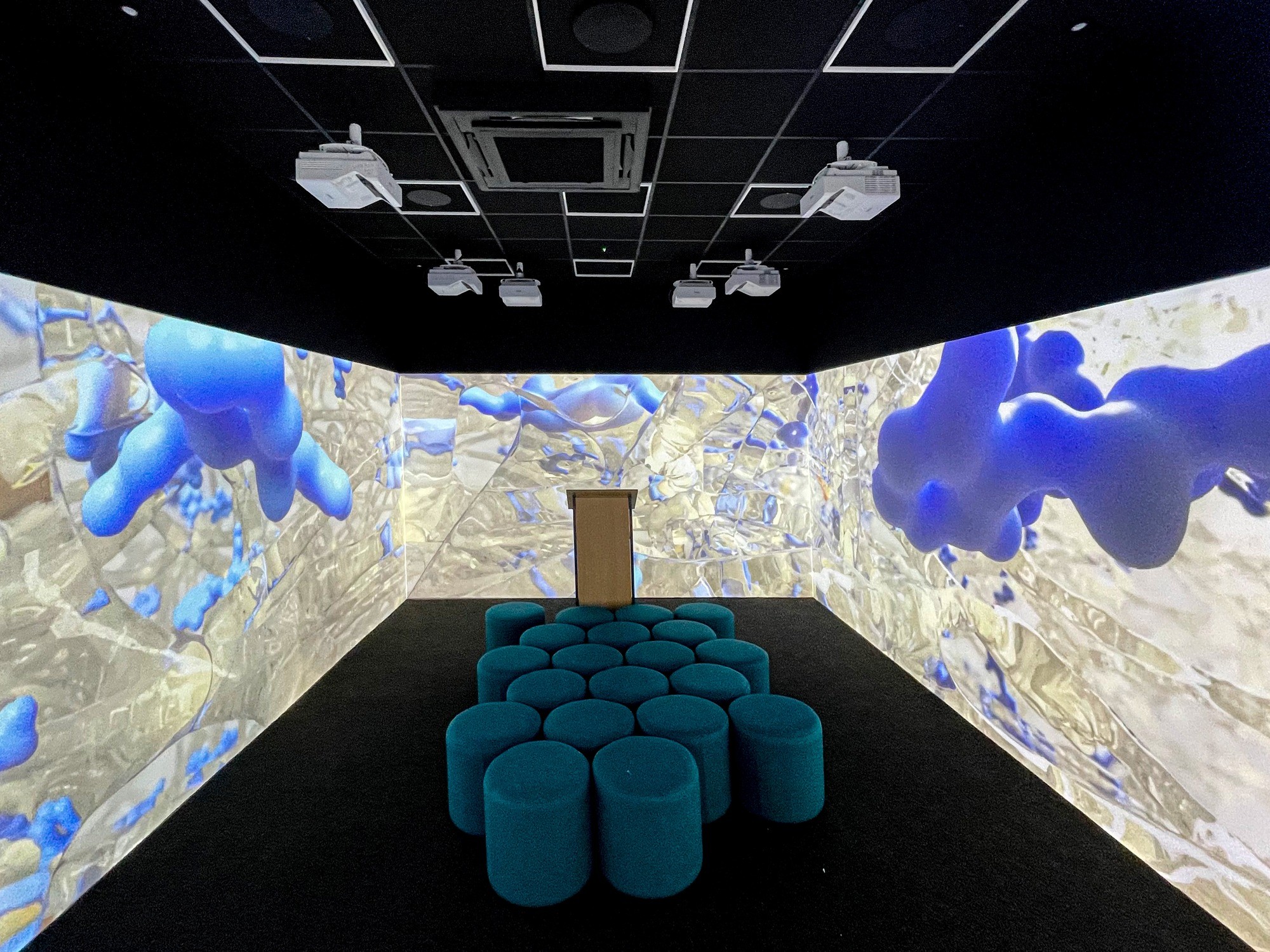 A futuristic room with wrapped projection screens displaying abstract blue and white shapes. A podium stands in front of teal circular stools.