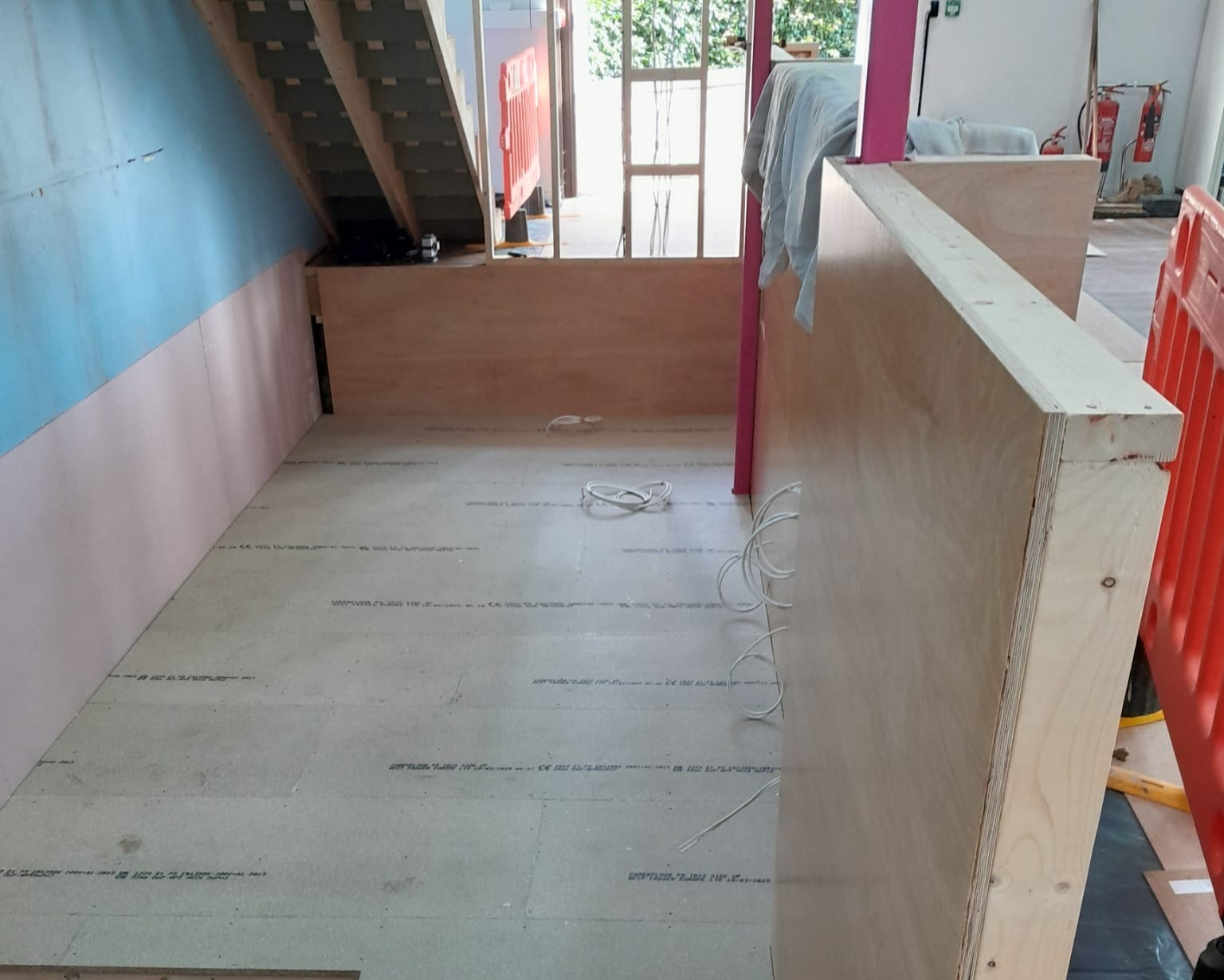 Construction area under a staircase with unfinished wooden walls and exposed wires on the floor. A blue wall on the left and bright orange barriers create a work-in-progress scene.