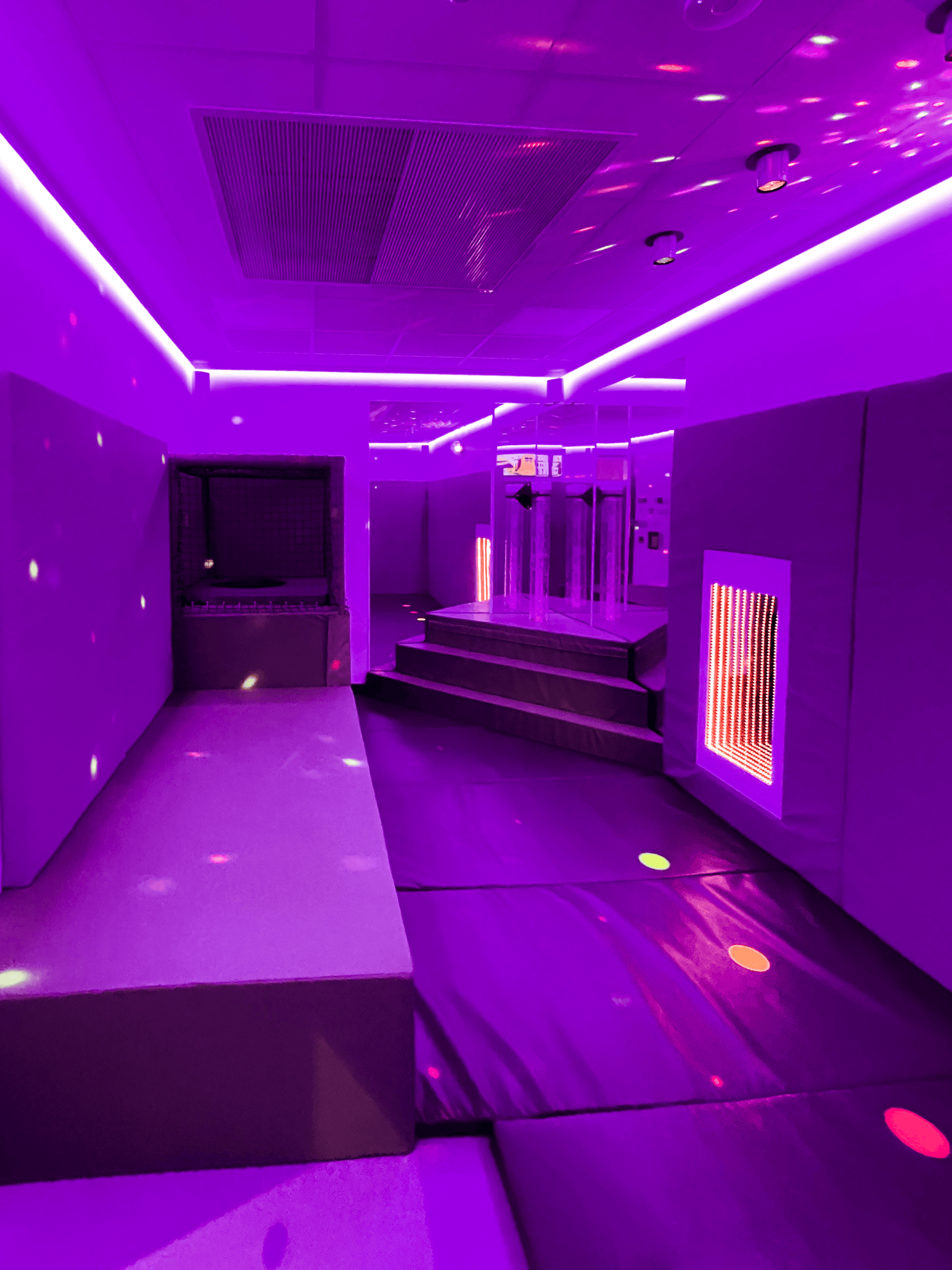 A softly lit room with purple LED lights, featuring cushioned platforms, a mirrored wall, and gentle sensory patterns. The atmosphere is calming and immersive.