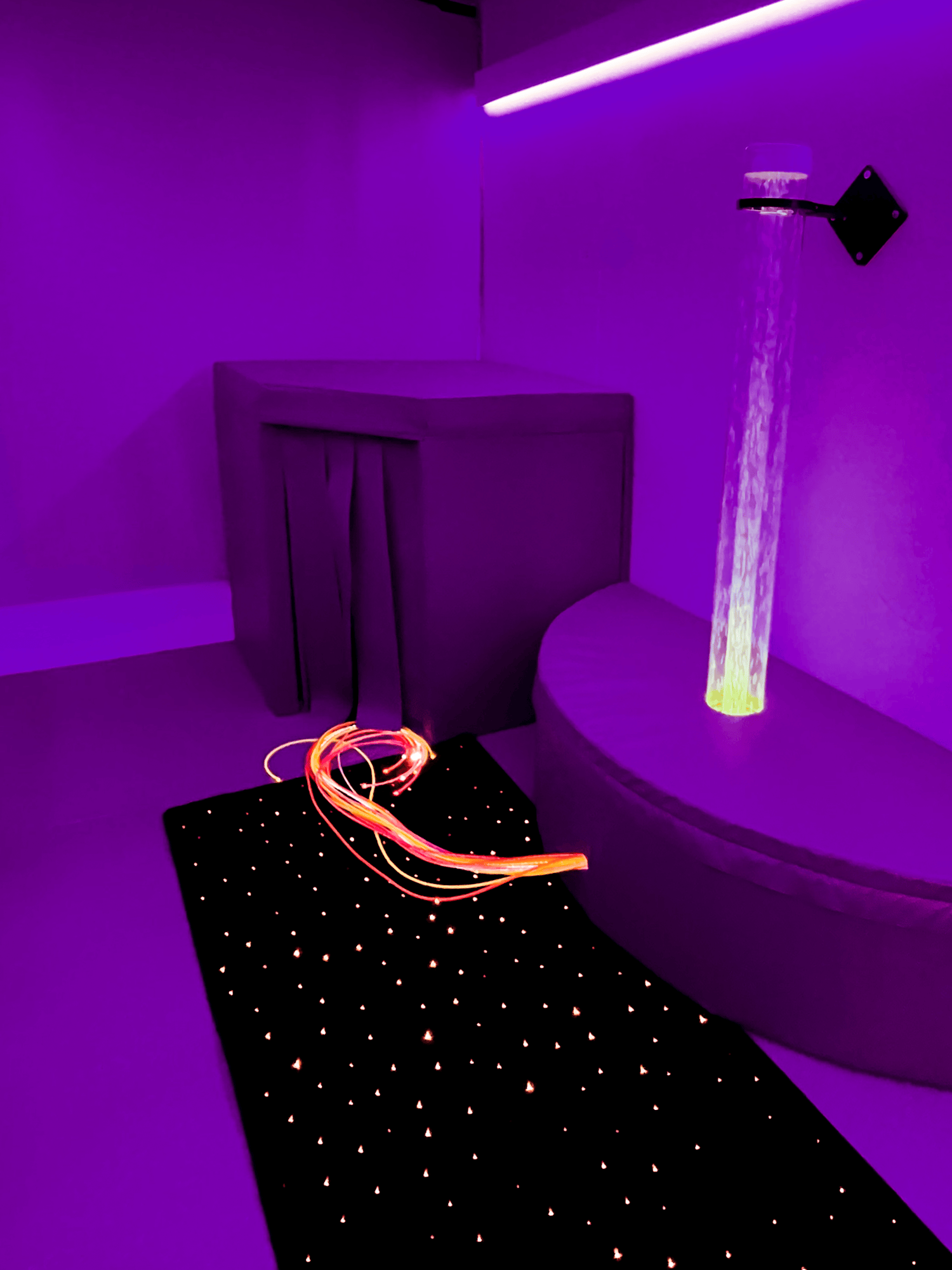Aurora installed room bathed in purple light features sensory objects: a glowing water tube, a fiber optic bundle, and a soft mat with tiny lights, creating a calming atmosphere.