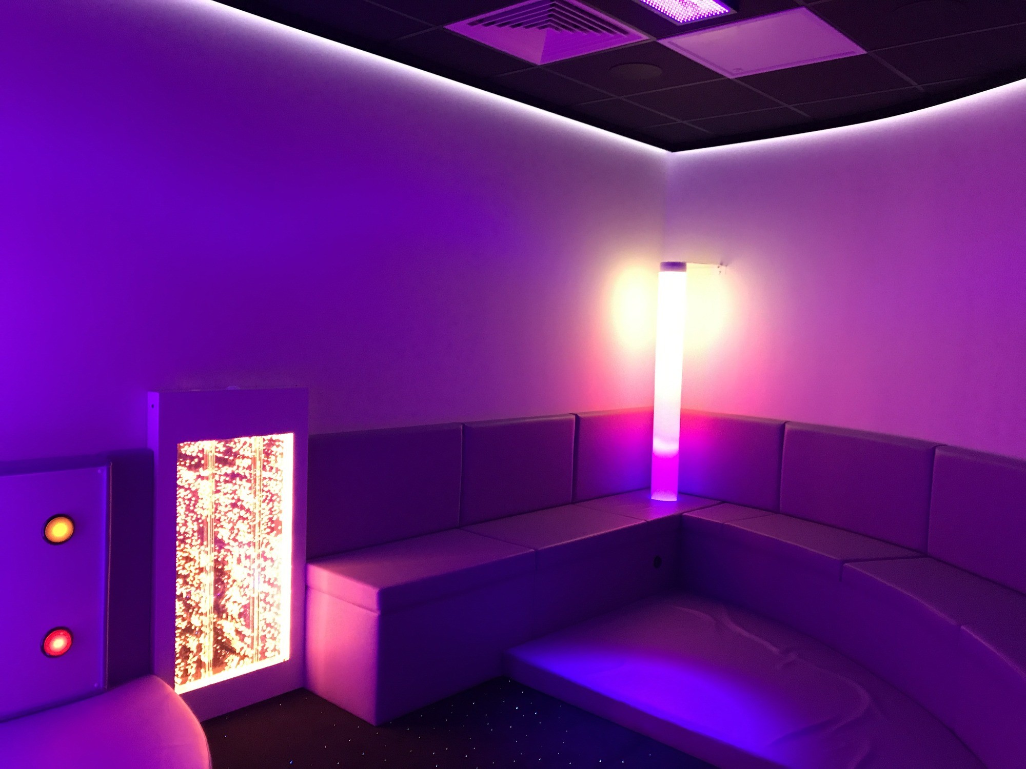 A softly lit sensory room features a curved purple couch, glowing bubble tubes, and ambient purple and yellow lighting, creating a calming atmosphere.
