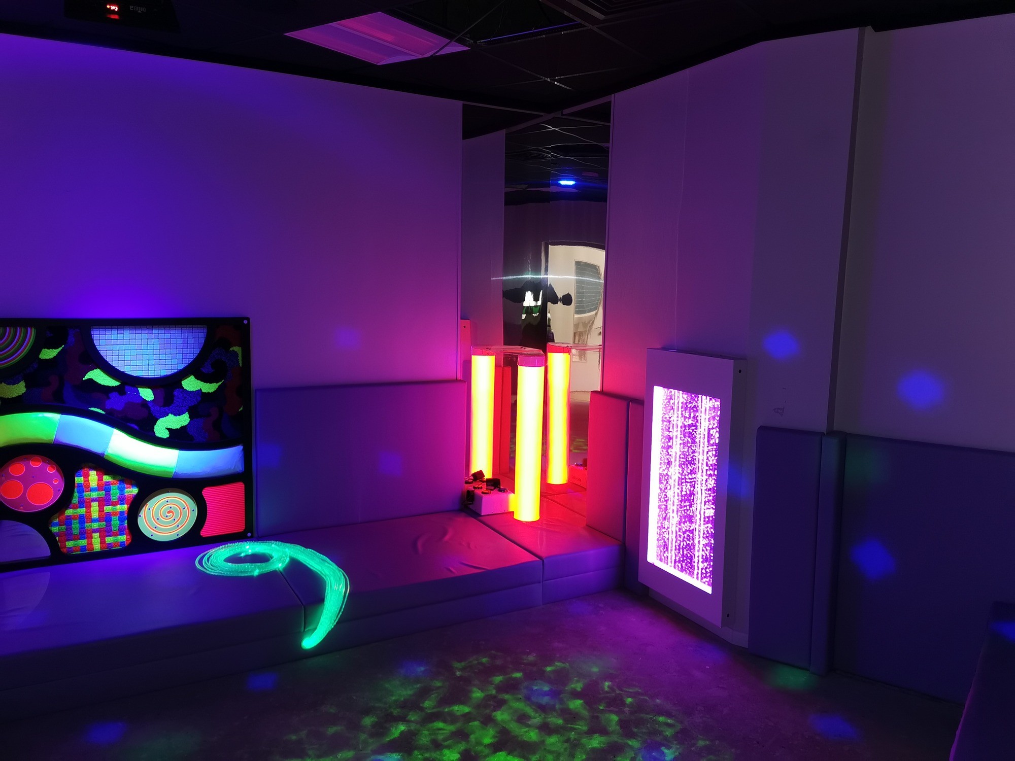 A sensory room with colorful neon lights: swirling patterns on a wall panel, glowing tubes, and illuminated bubble columns create a calming atmosphere.