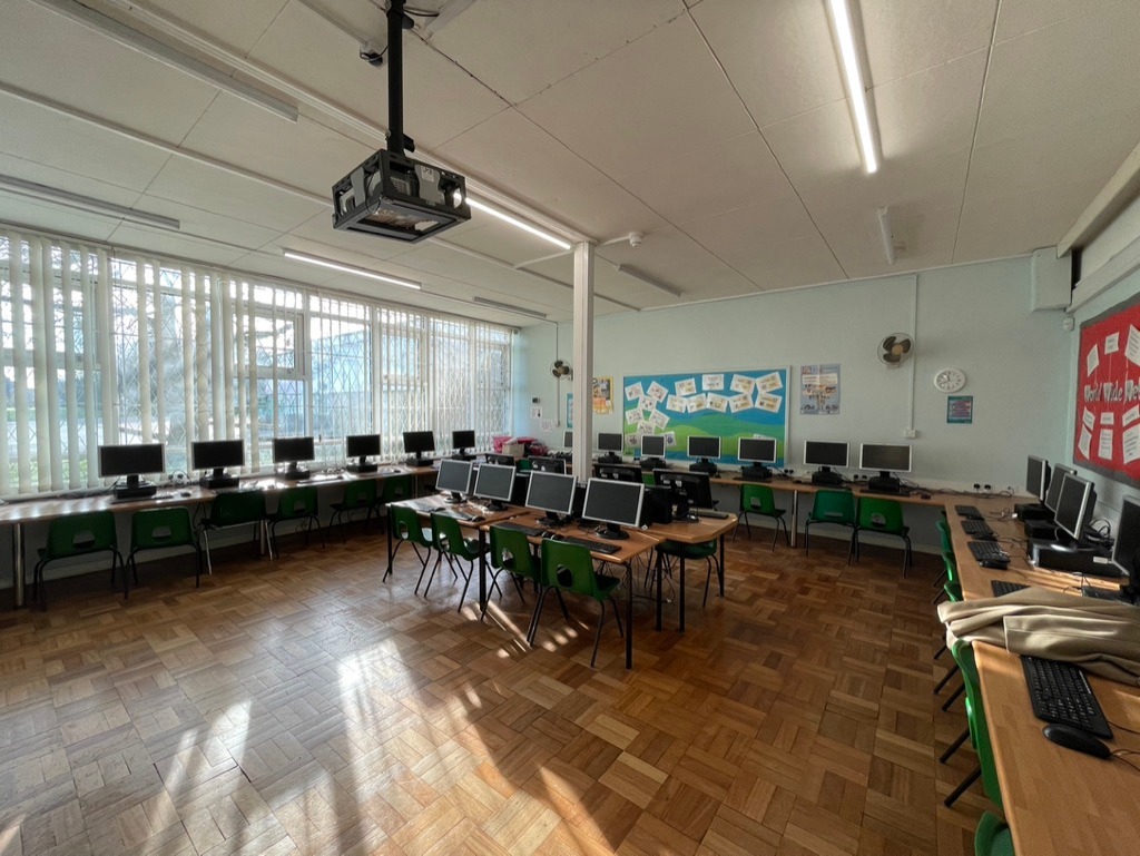 Spacious classroom with parquet flooring, lined with rows of computers on wooden desks. Large windows let in natural light, creating a bright atmosphere.