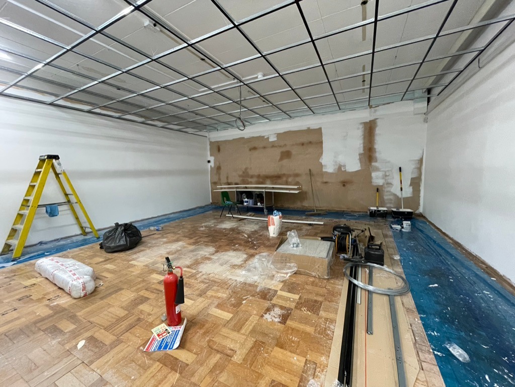 Room under renovation with exposed ceiling grid and unfinished walls. Wooden floor has scattered tools and materials. Yellow ladder leans on left wall.