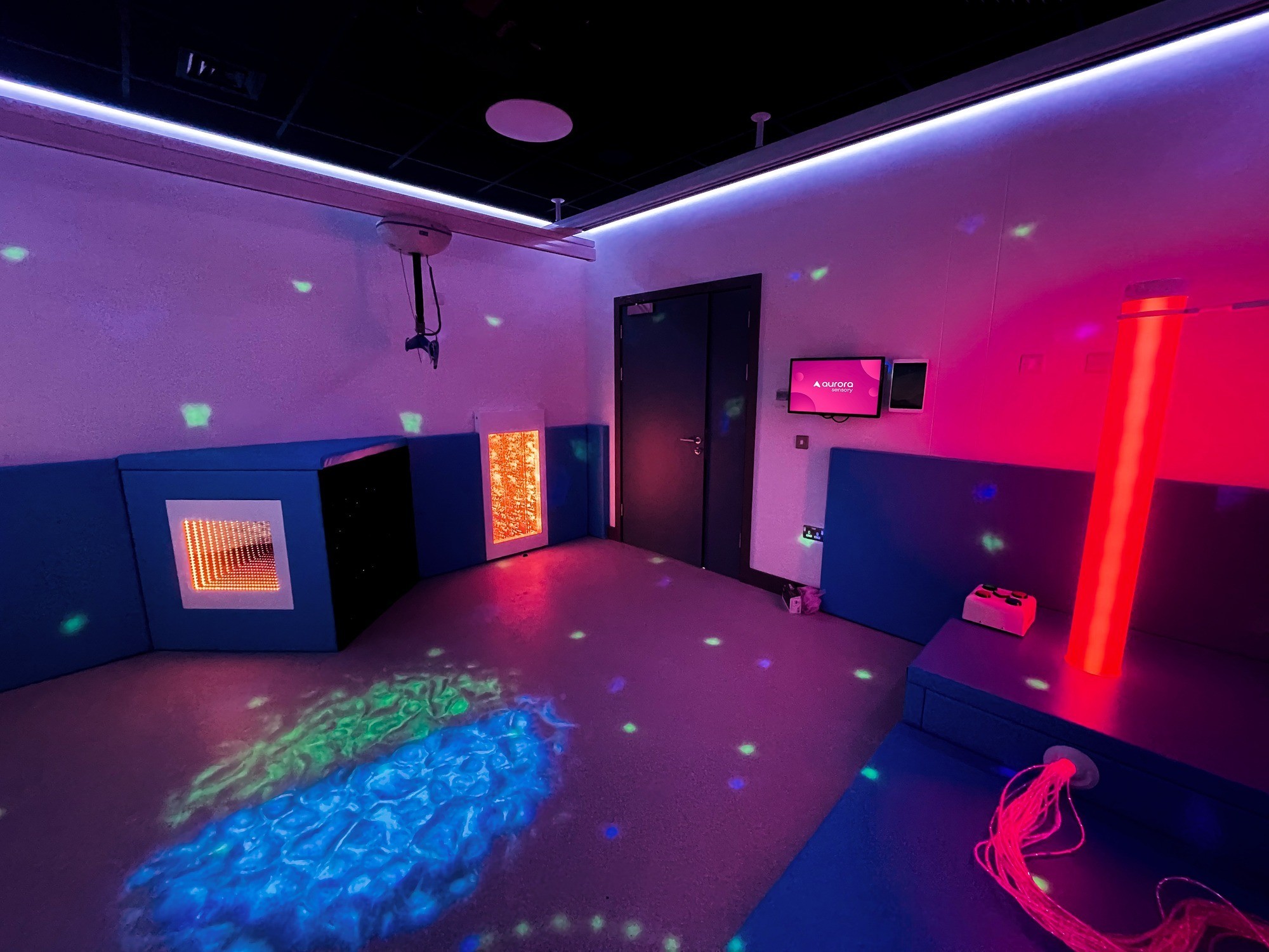 A sensory room with soft, colorful lighting. Features include a glowing bubble tube, fiber optic strands, and a shimmering wall panel, creating a calming atmosphere.