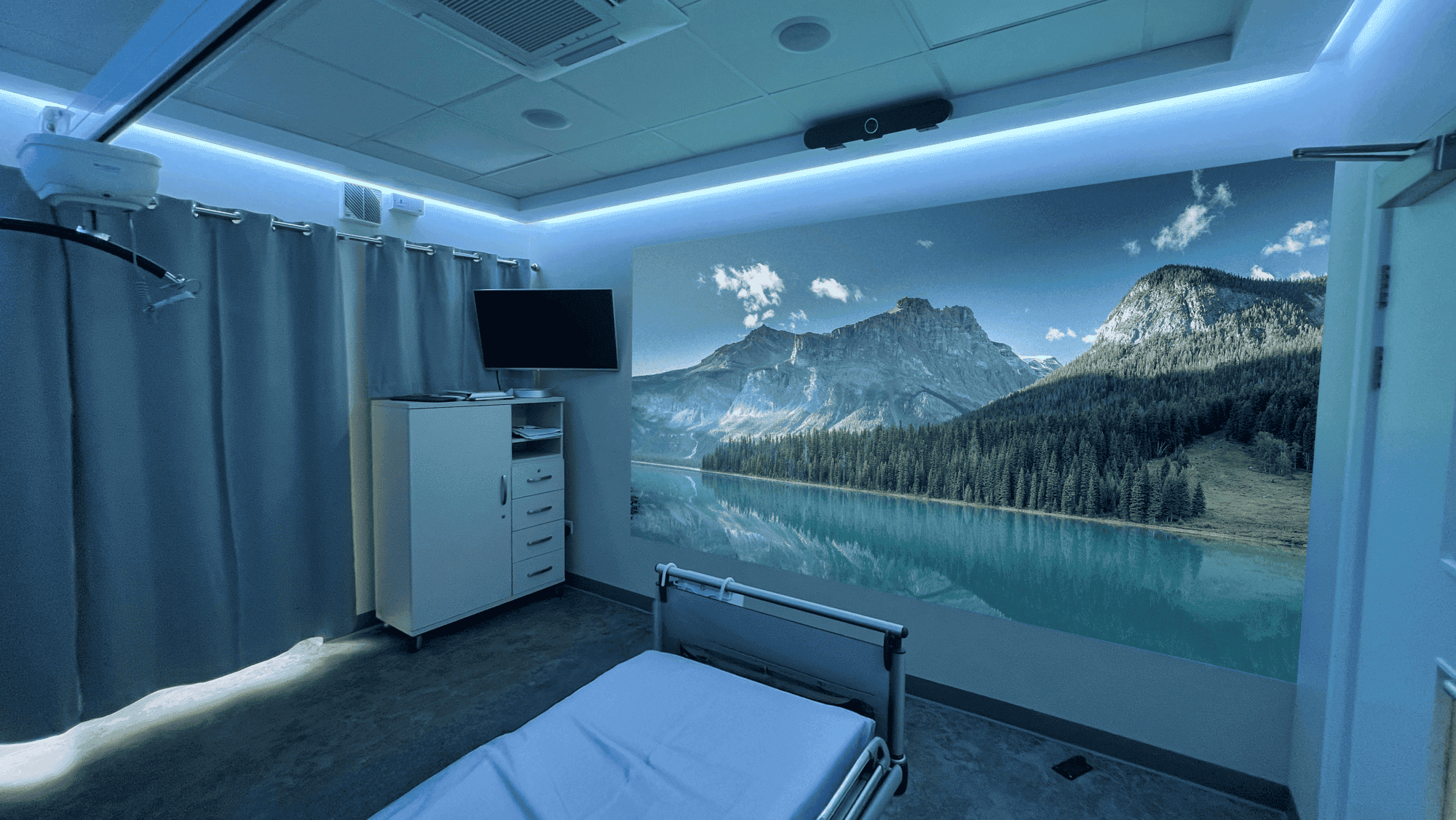 Hospital room with a white bed, grey curtains, hidden ambient coloured lighting and a single wall aurora projection image of a nature mural of mountains and a lake creates a calming atmosphere.
