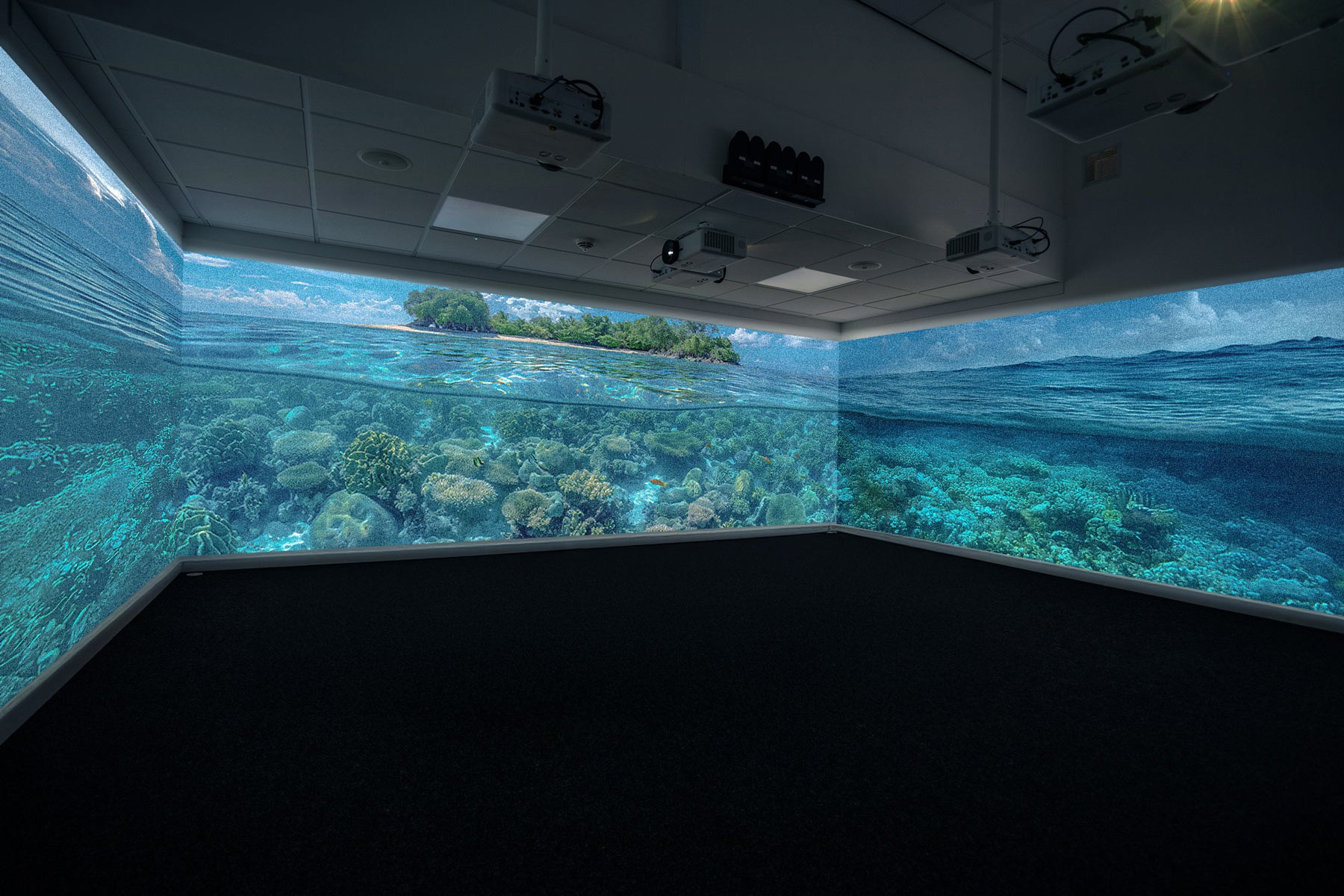 A dark room with three wall projection displaying a vivid underwater scene features coral reefs, clear blue water, and an island horizon, creating an immersive aquatic experience.