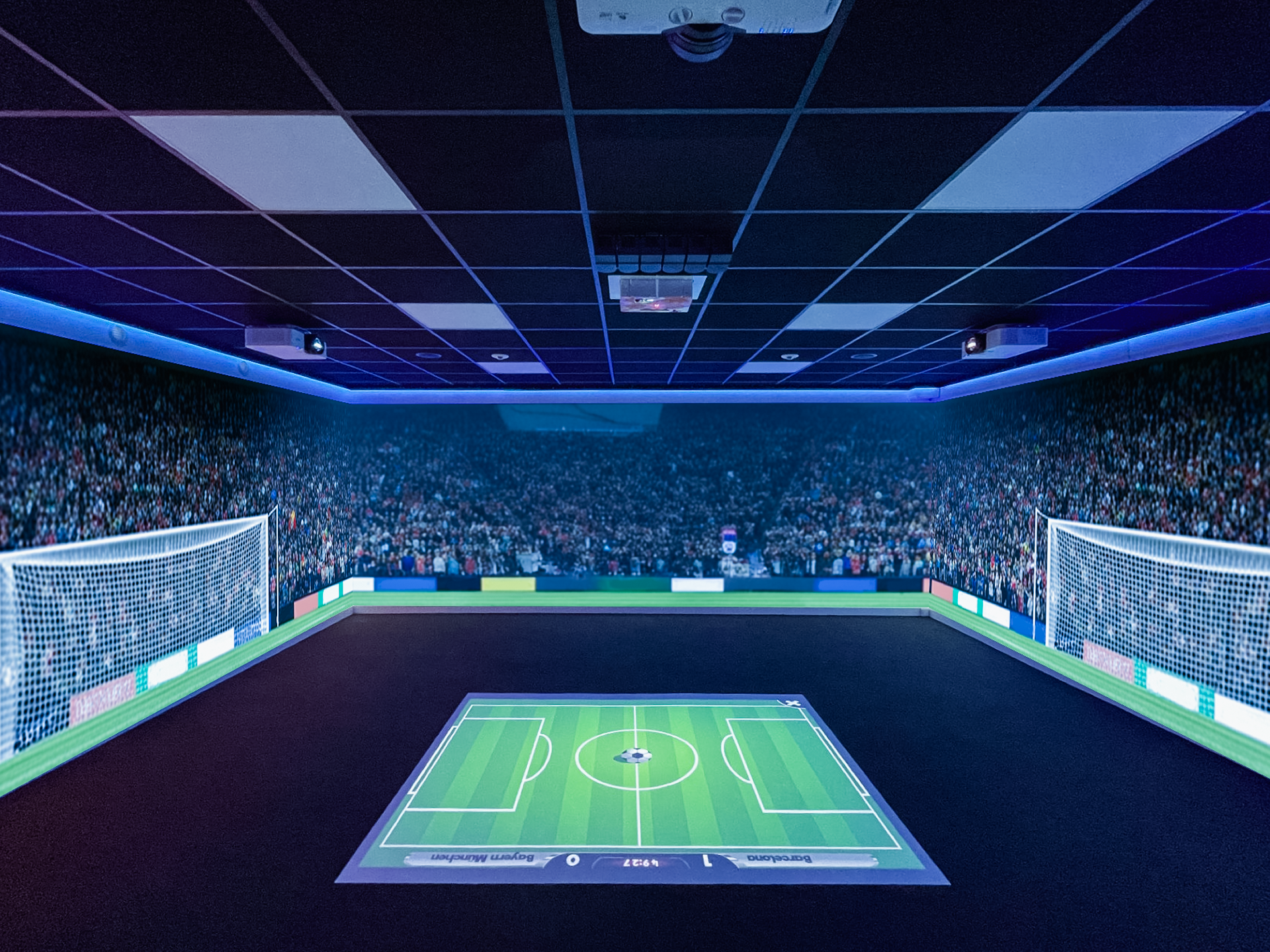 Indoor room designed as a soccer stadium with projected field on floor, digital screens displaying a cheering crowd, and goals on both ends.