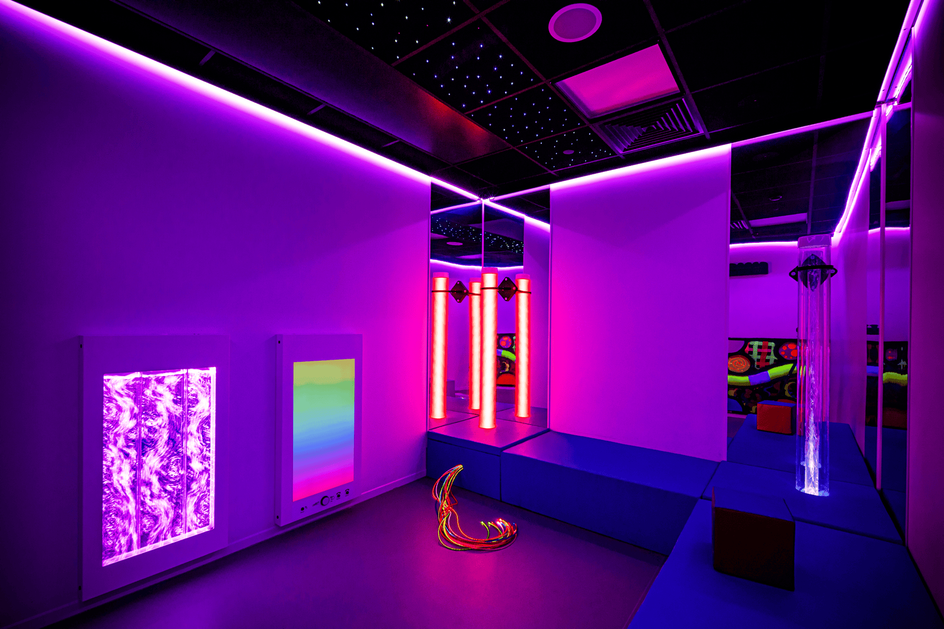 Darkened sensory environment with seating. Lit by bright pink and purple led lights, vortex panels on the wal, fibre optic cables and chroma tubes.